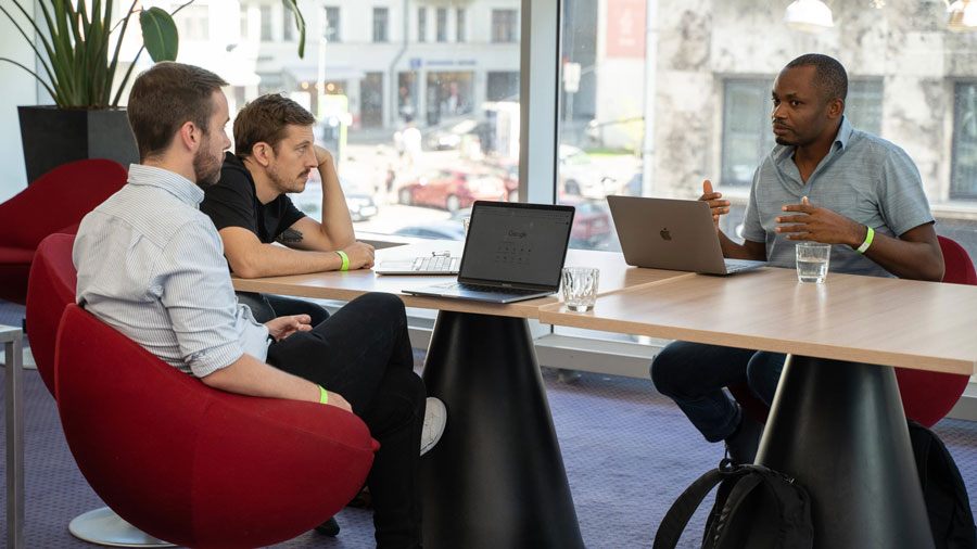 Four people sitting at a table with laptops, engaged in a discussion in a brightly lit room with a large window.